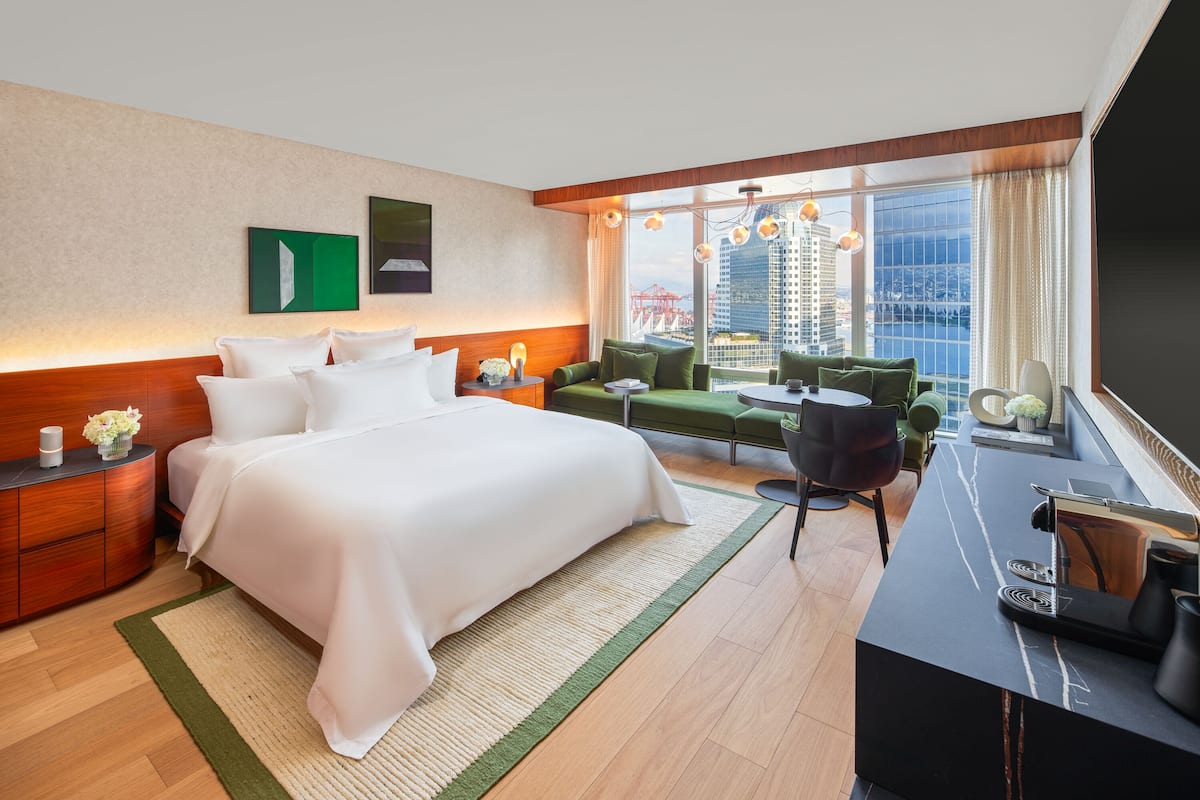 A modern hotel room with a large bed, a desk, a sofa, and a view of a cityscape.