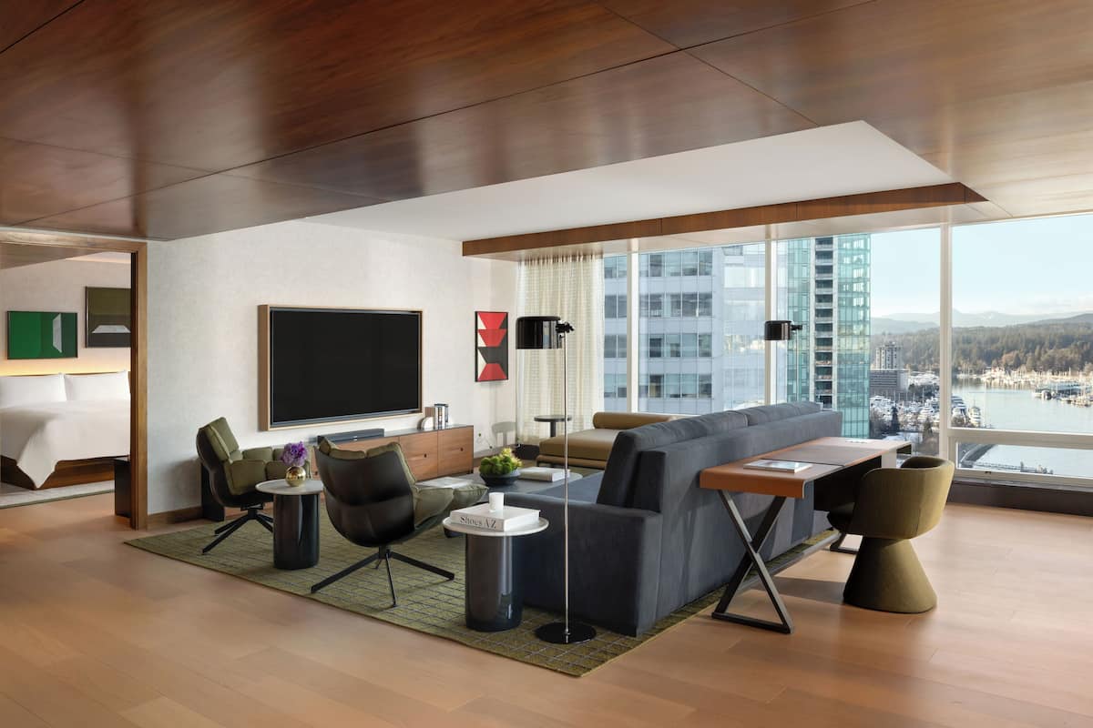 A modern hotel room with a large sofa, a dining table, and a flat-screen TV. The room has large windows offering a view of the city and a body of water.