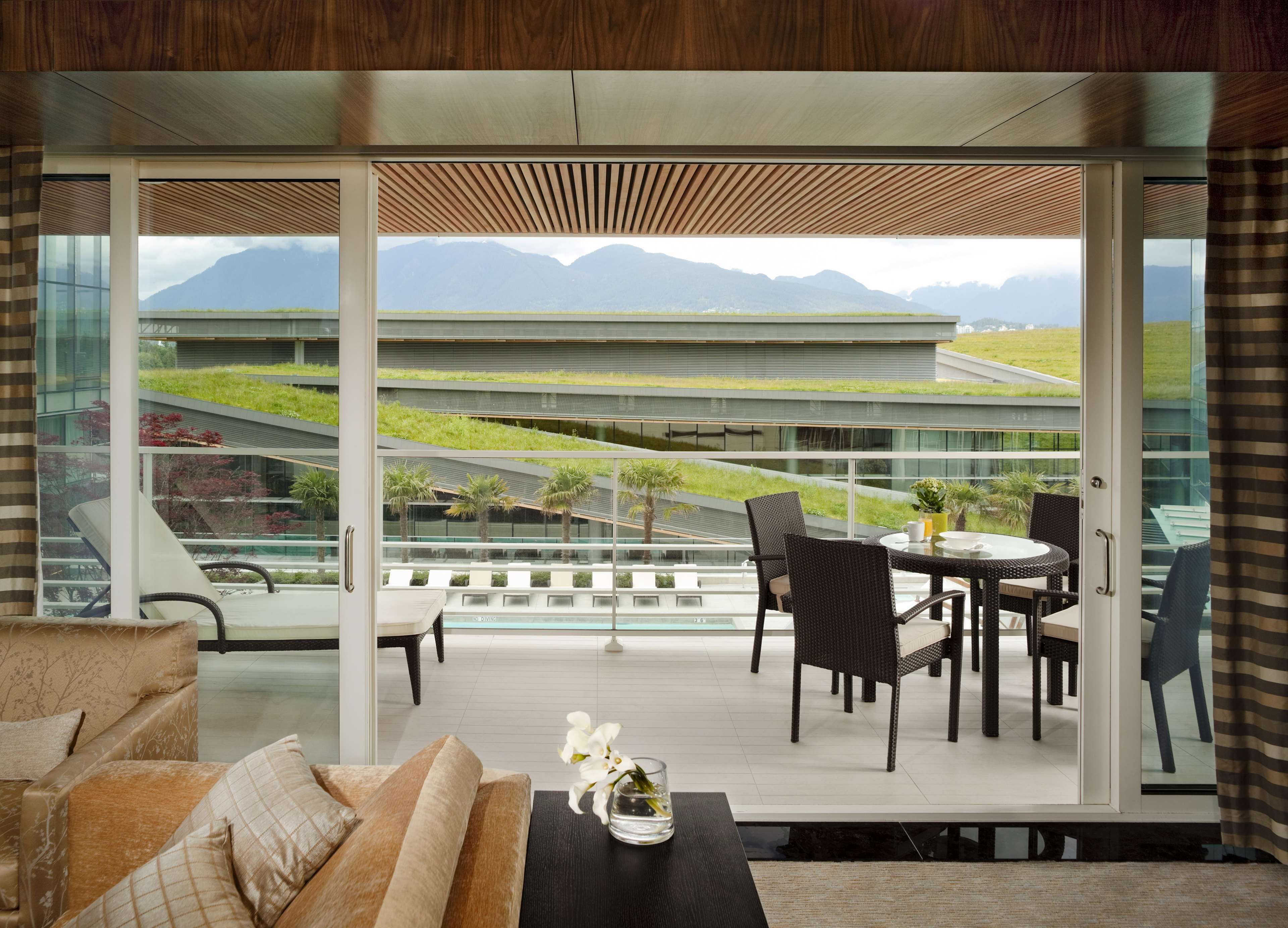 Ein modernes Wohnzimmer mit einem großen Schiebeglastür, die auf einen Balkon mit Blick auf ein Gebäude mit begrünten Dächern und Berge im Hintergrund führt.