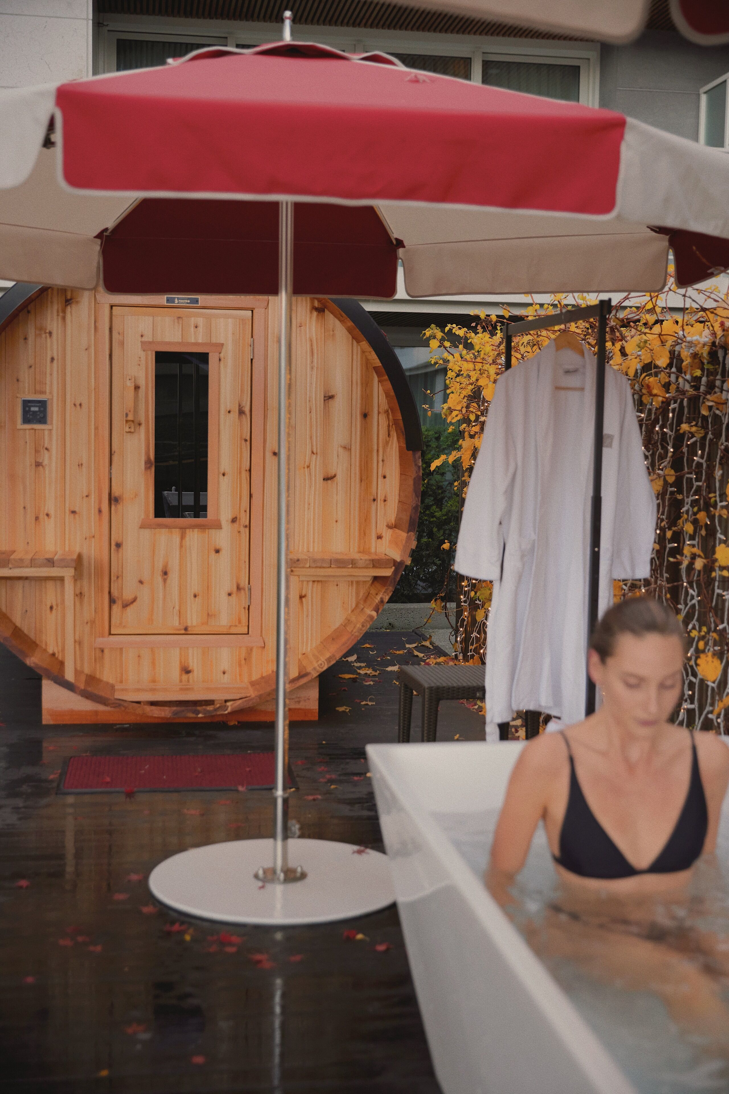 A wooden sauna with a red umbrella, a white robe hanging, and a person in a bathtub.