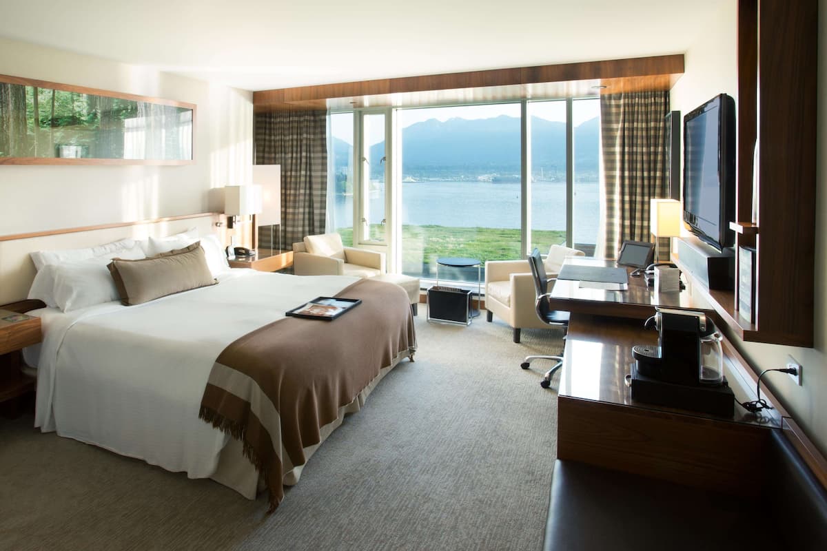 A hotel room with a large bed, a desk with a coffee maker, a TV, and a view of a lake and mountains.