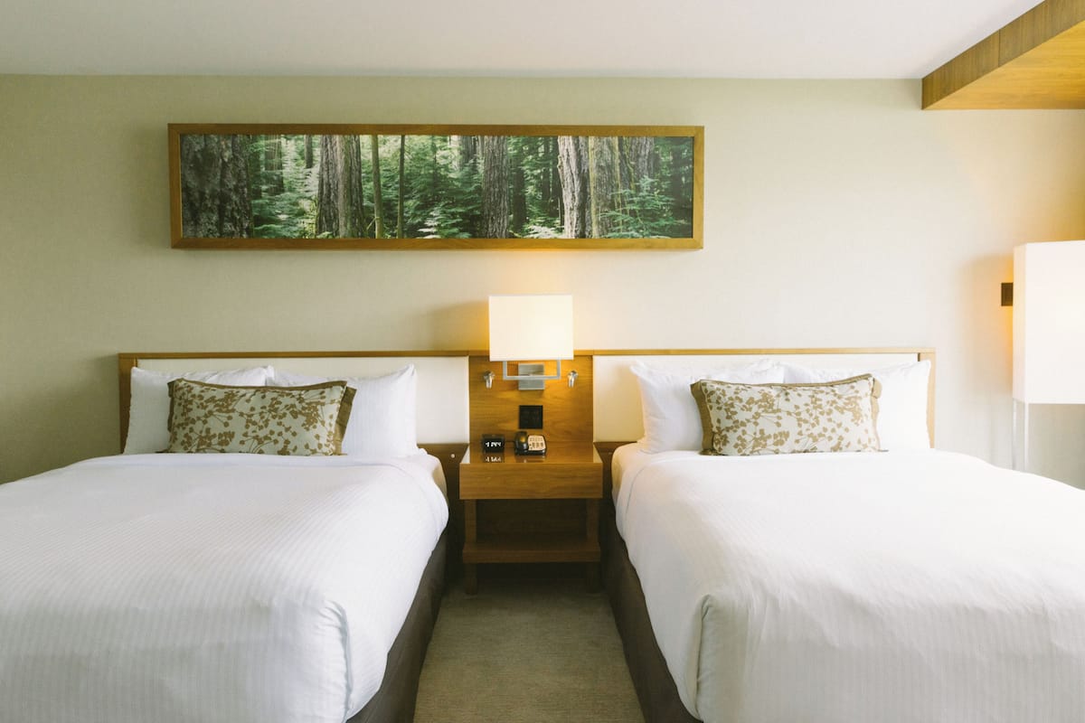 A hotel room with two beds, a nightstand with a lamp, a painting of a forest, and a bedside table with a clock and a phone.