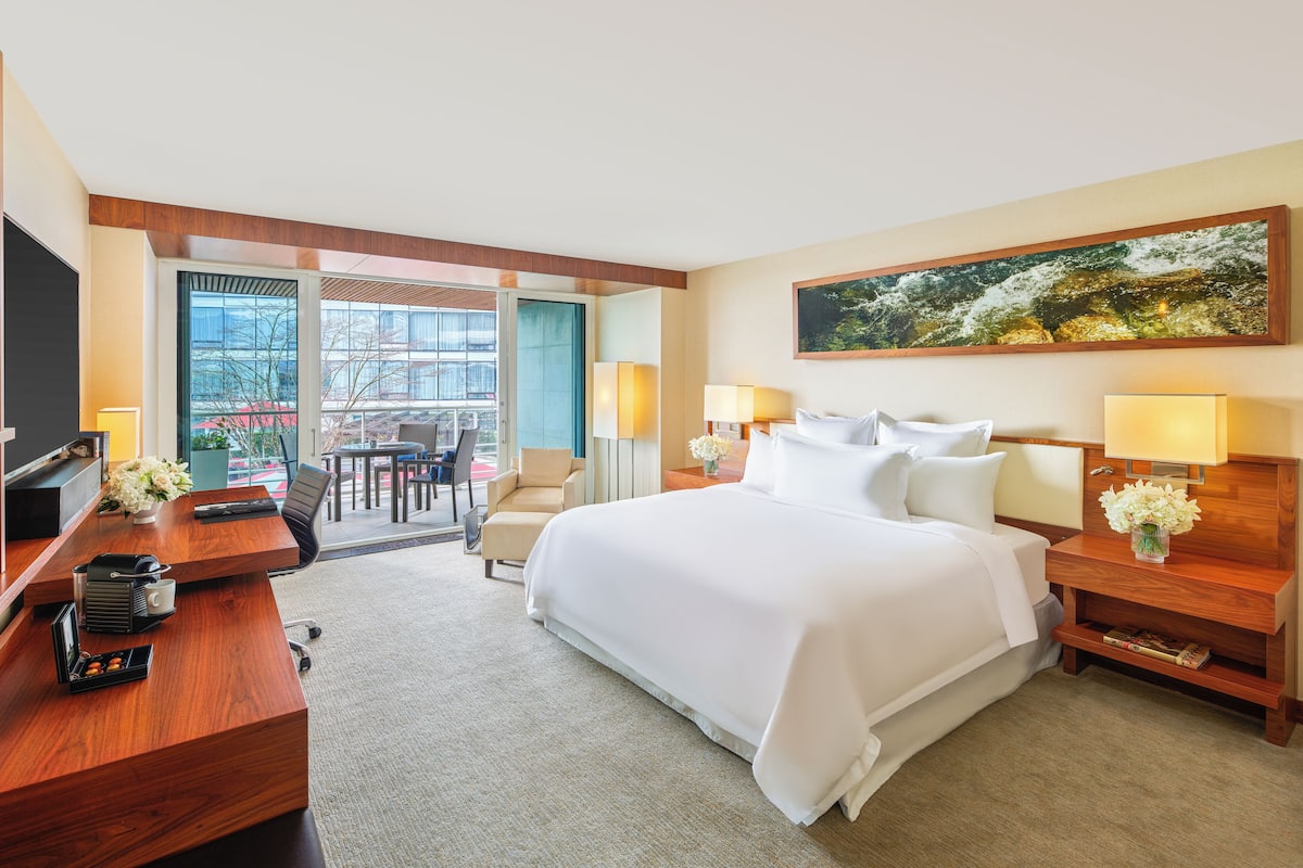 A hotel room with a large bed, a desk with a computer, a chair, a small table with flowers, and a view of the outdoors.