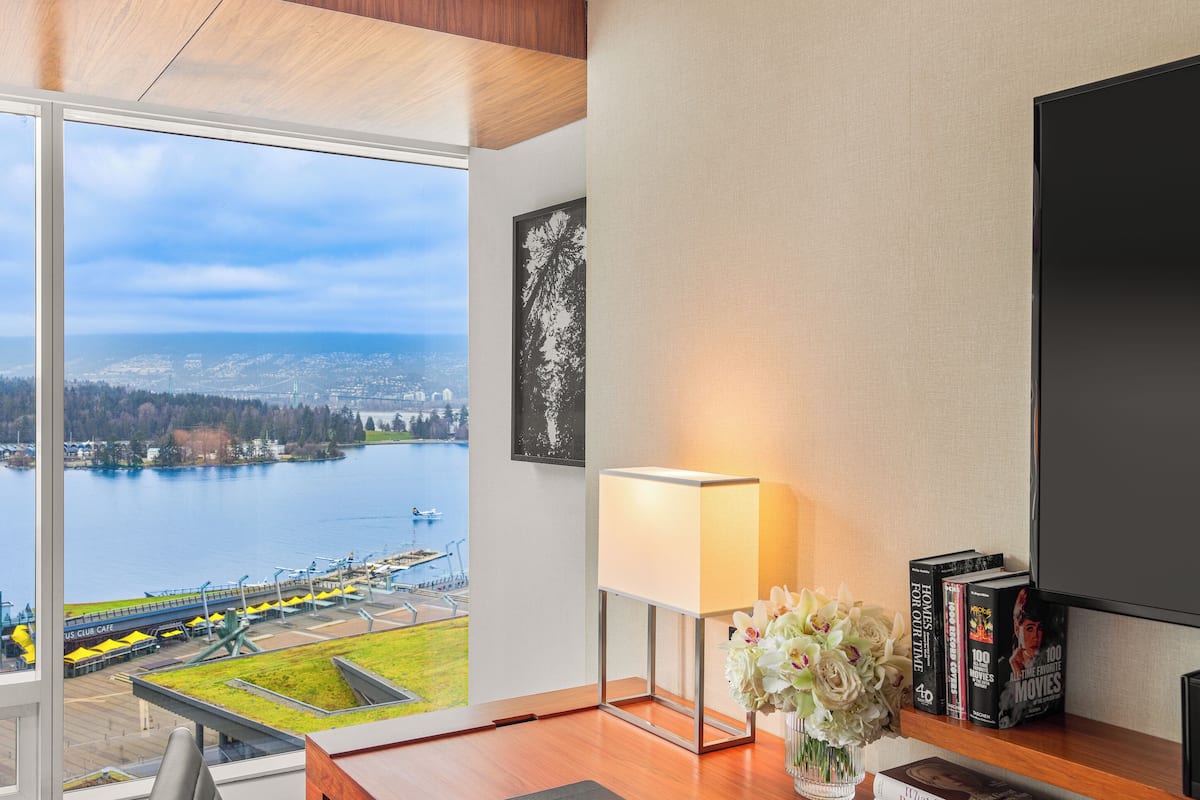 A hotel room with a wooden desk, a chair, a lamp, a vase with flowers, a bookshelf with books, a flat-screen TV, and a large window with a view of a body of water and a cityscape.