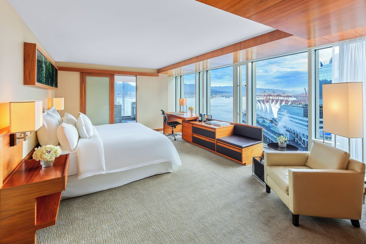 A modern hotel room with a large bed, a desk, a sofa, and a view of a cityscape through large windows.