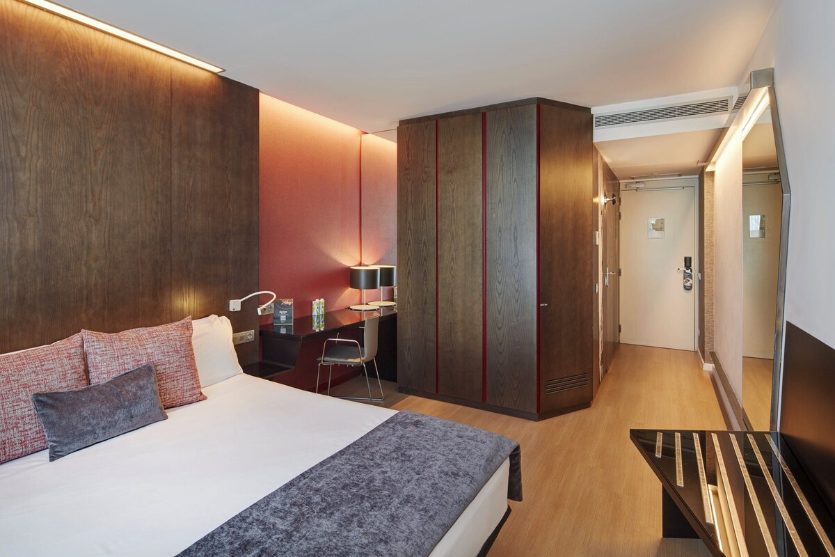 A modern hotel room with a large bed, a desk, and a wardrobe.