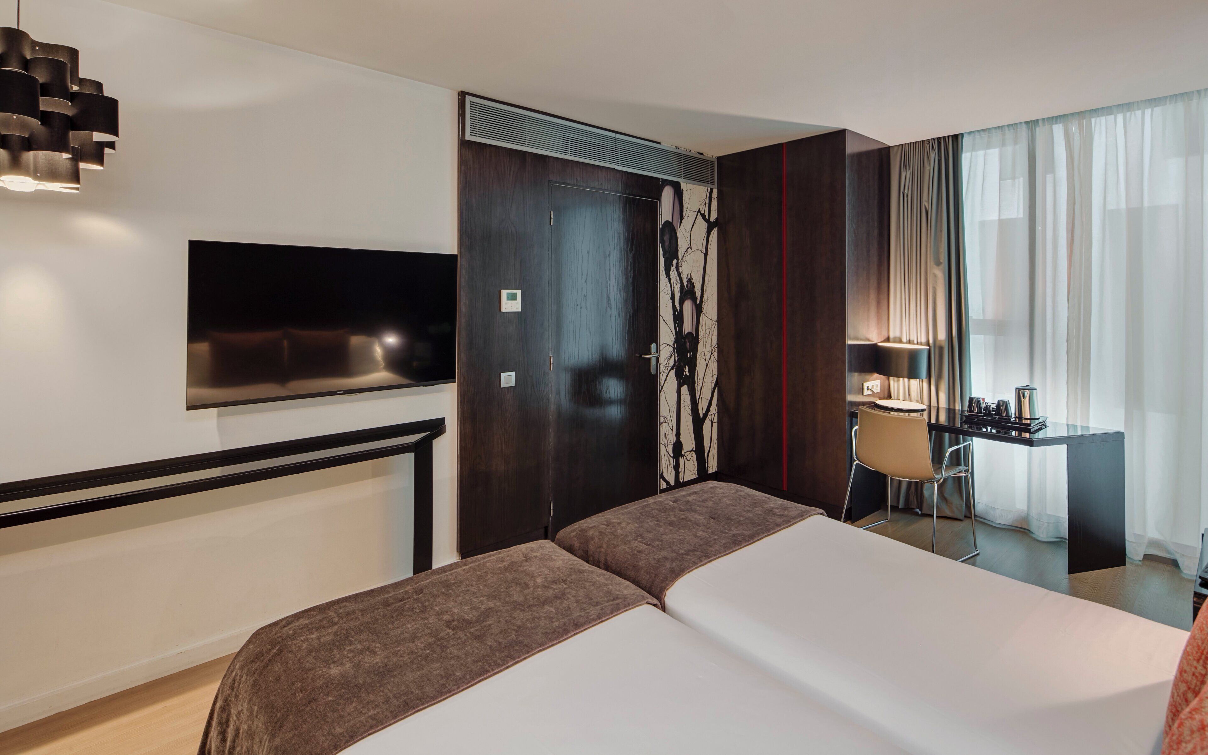 A hotel room with a large bed, a flat-screen TV mounted on the wall, a desk with a chair, and a window with sheer curtains.