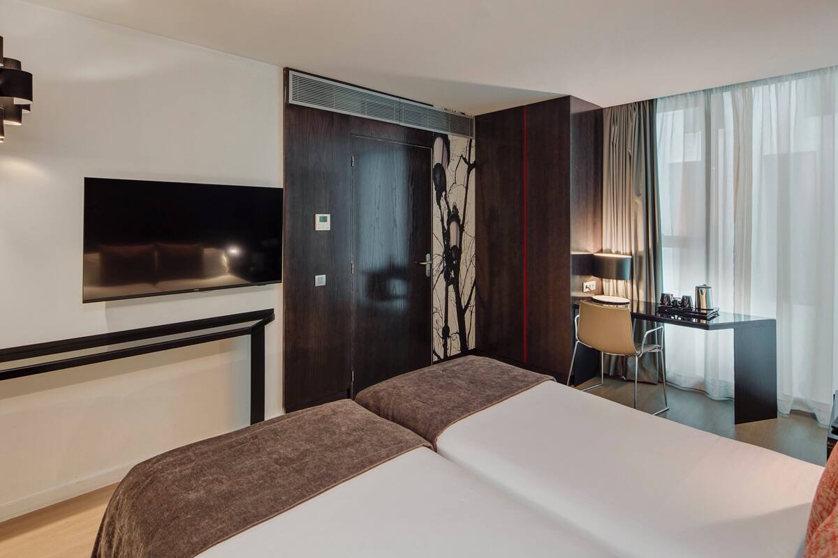 Une chambre d’hôtel dotée d’un grand lit, d’un téléviseur à écran plat fixé au mur, d’un bureau avec une chaise et d’une fenêtre avec des rideaux légers.