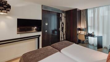 A hotel room with a large bed, a flat-screen TV mounted on the wall, a desk with a chair, and a window with sheer curtains.