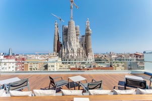 A rooftop terrace with wooden flooring, white cushioned chairs, and a table. A large building with a unique architectural design and construction cranes is visible in the background.