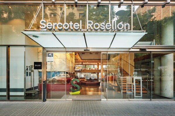 Mercotel Rosellón -hotellin sisäänkäynti, jossa on suuri lasijulkisivu, selkeästi näkyvä vastaanottoalue ja portaikko, joka johtaa ylemmissä kerroksiin.