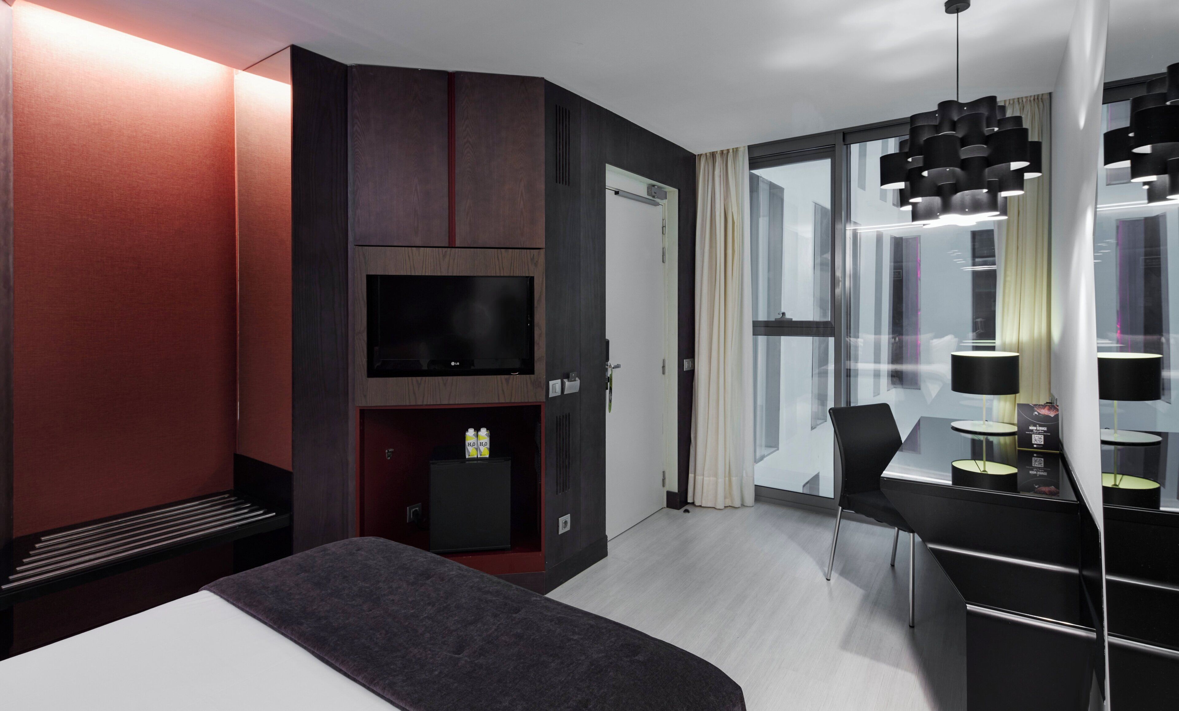 Ett modernt hotellrum med en säng, en TV, ett skrivbord med en stol och ett stort fönster.