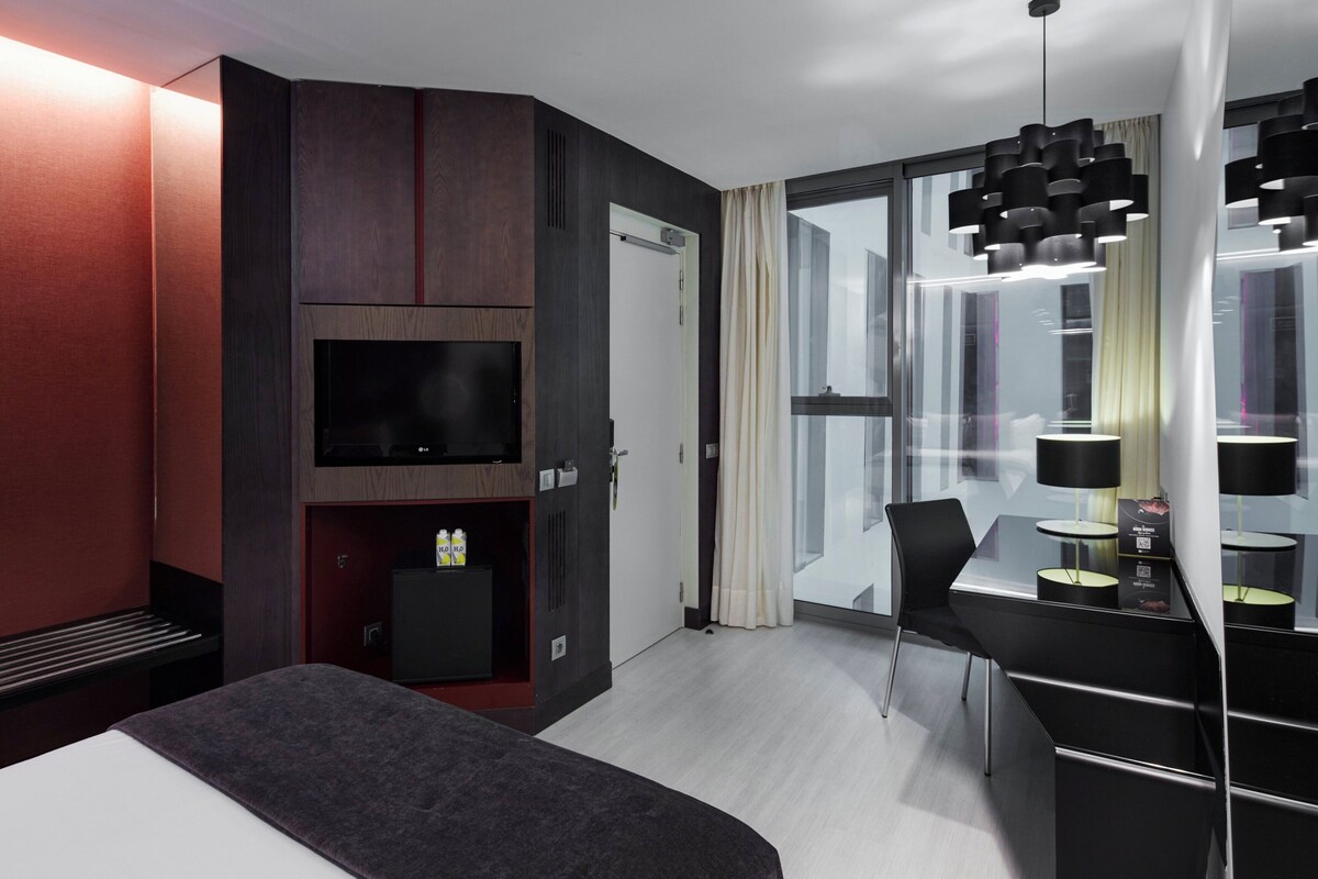A modern hotel room with a bed, a TV, a desk with a chair, and a large window.