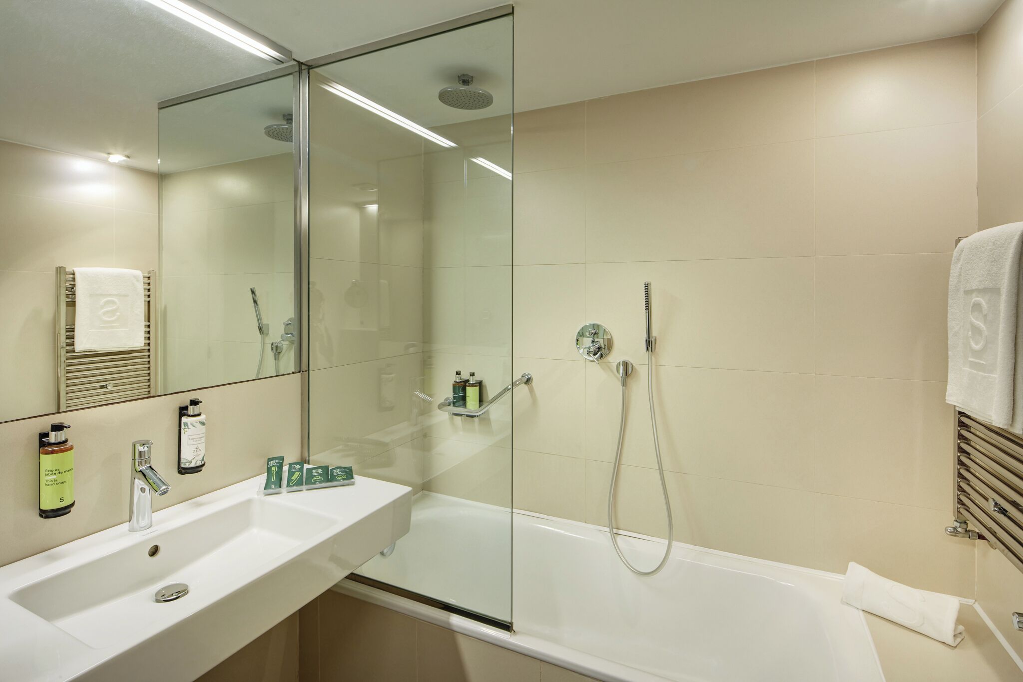 Un baño moderno con ducha de vidrio, lavabo blanco con artículos de aseo y un espejo grande.