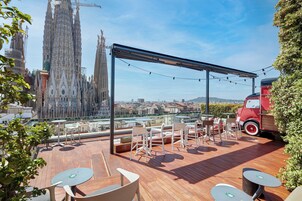 Terraço no telhado com piso de madeira, cadeiras e mesas brancas, e um veículo vermelho.