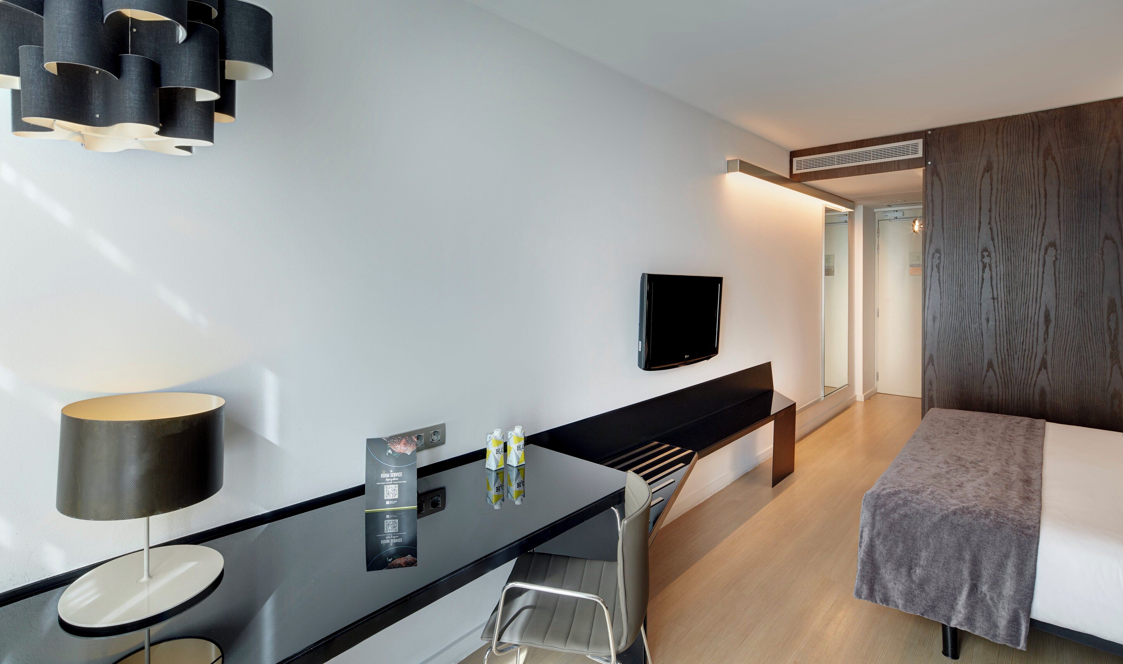 Habitación de hotel moderna con una televisión de pantalla plana montada en la pared, un escritorio con lámpara y una cama con manta gris.