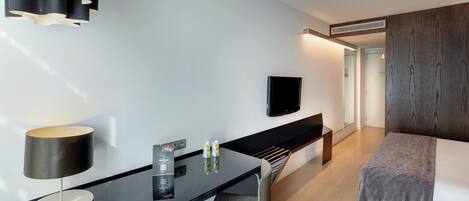 Habitación de hotel moderna con una televisión de pantalla plana montada en la pared, un escritorio con lámpara y una cama con manta gris.