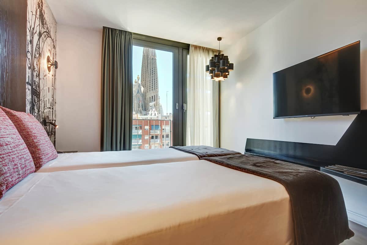 A hotel room with two beds, a large window with a view of a building, a flat-screen TV, and a modern pendant light.