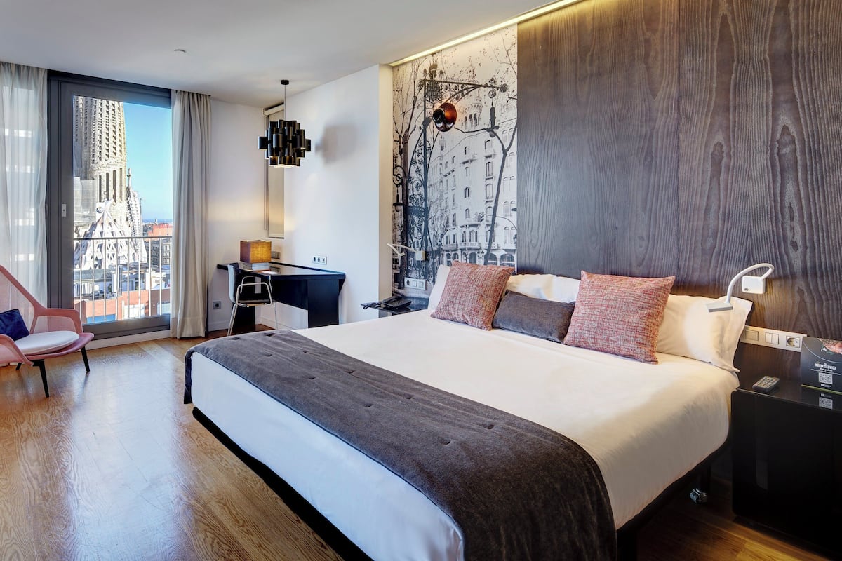 A modern hotel room with a large bed, a desk, a chair, and a view of a cityscape through the window.