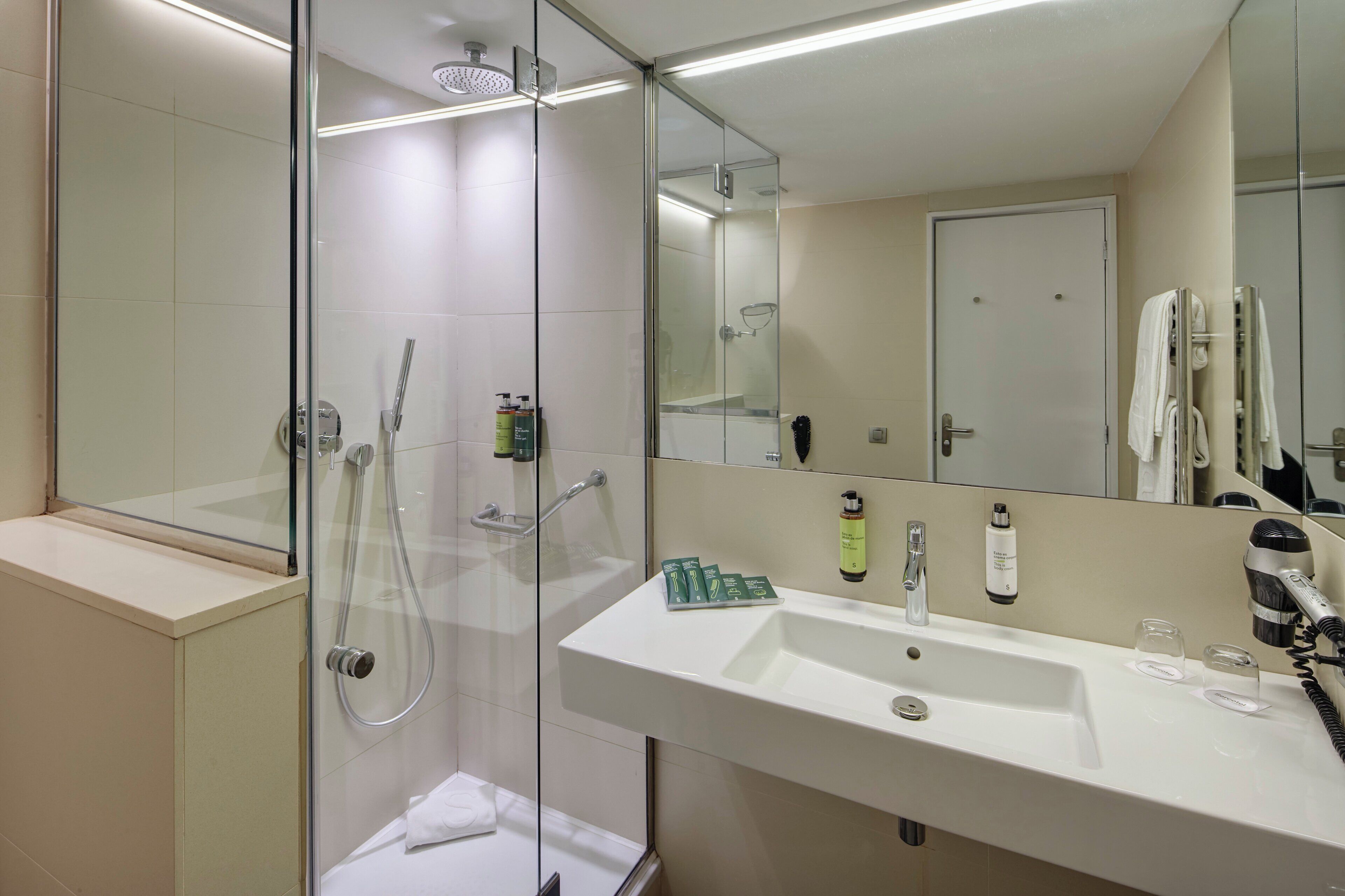 Un baño moderno con ducha envuelta en vidrio, un lavabo blanco y un espejo que refleja una puerta y toallas.