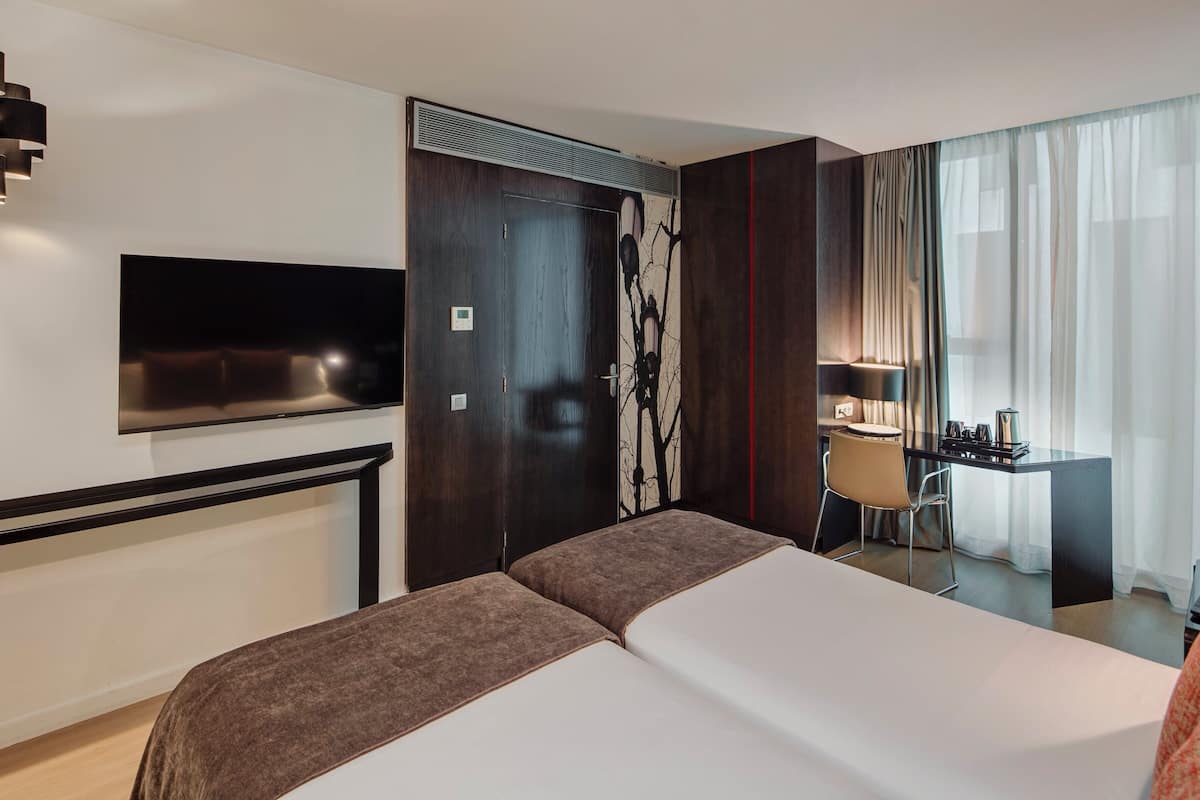 A hotel room with a large bed, a flat-screen TV mounted on the wall, a desk with a chair, and a window with sheer curtains.