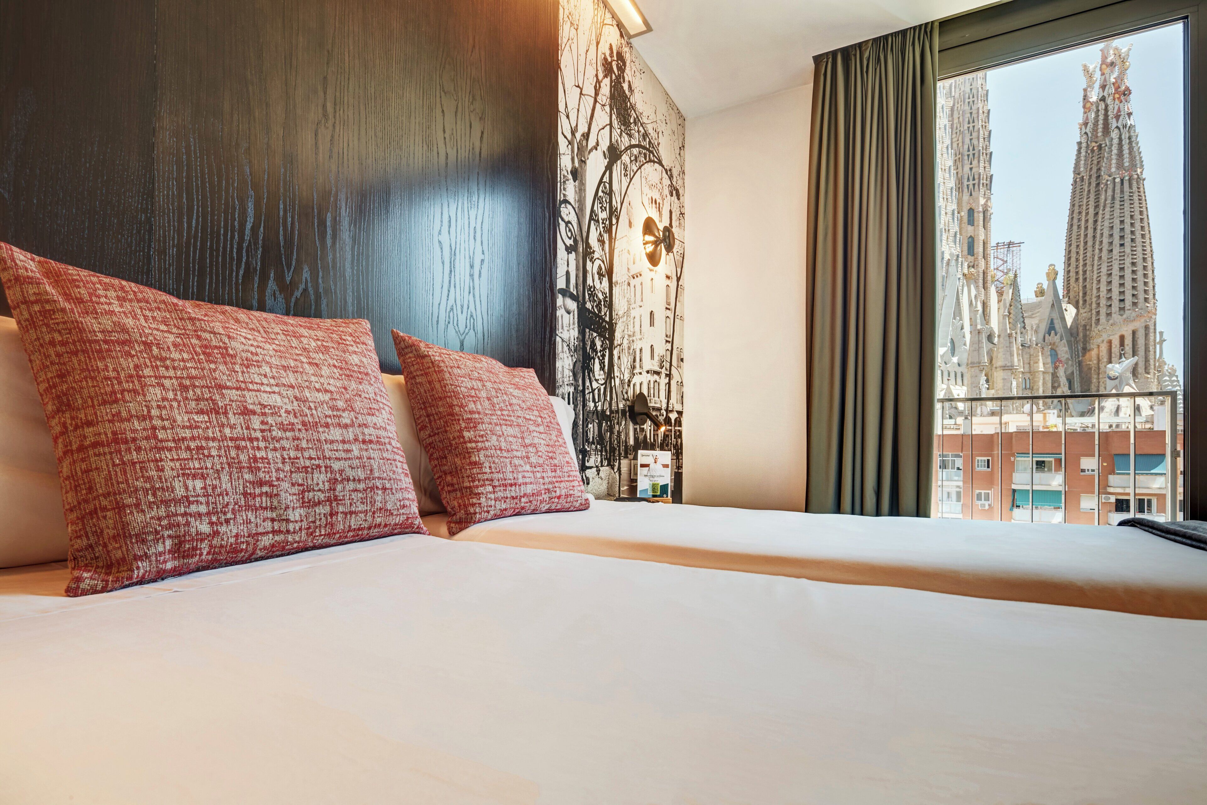 Habitación de hotel con una cama grande, almohadones estampados en rojo, cabecera de madera oscura, un mural de un paisaje urbano y vista a un edificio alto.