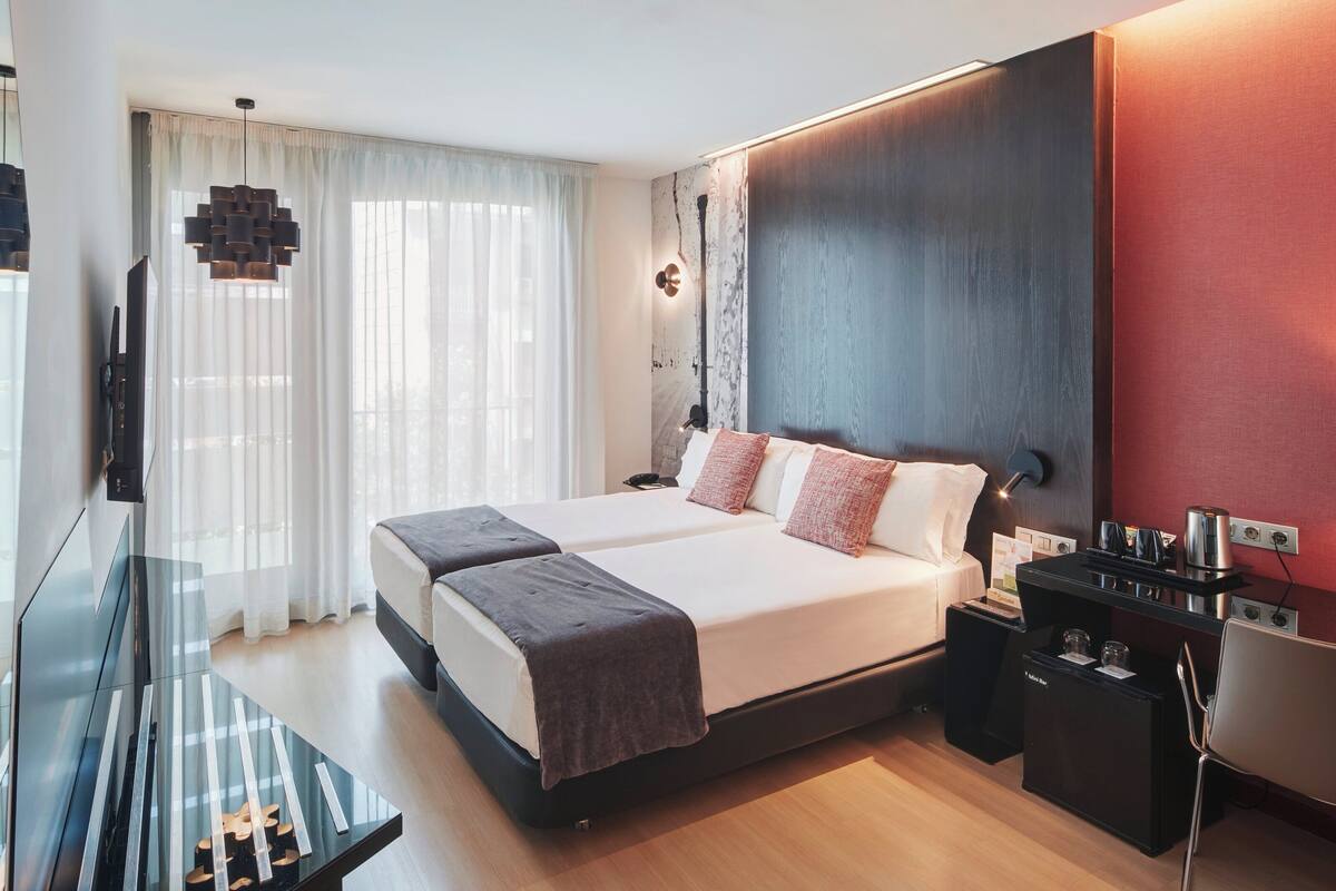A modern hotel room with a large bed, a desk with a chair, a TV, and a minibar.