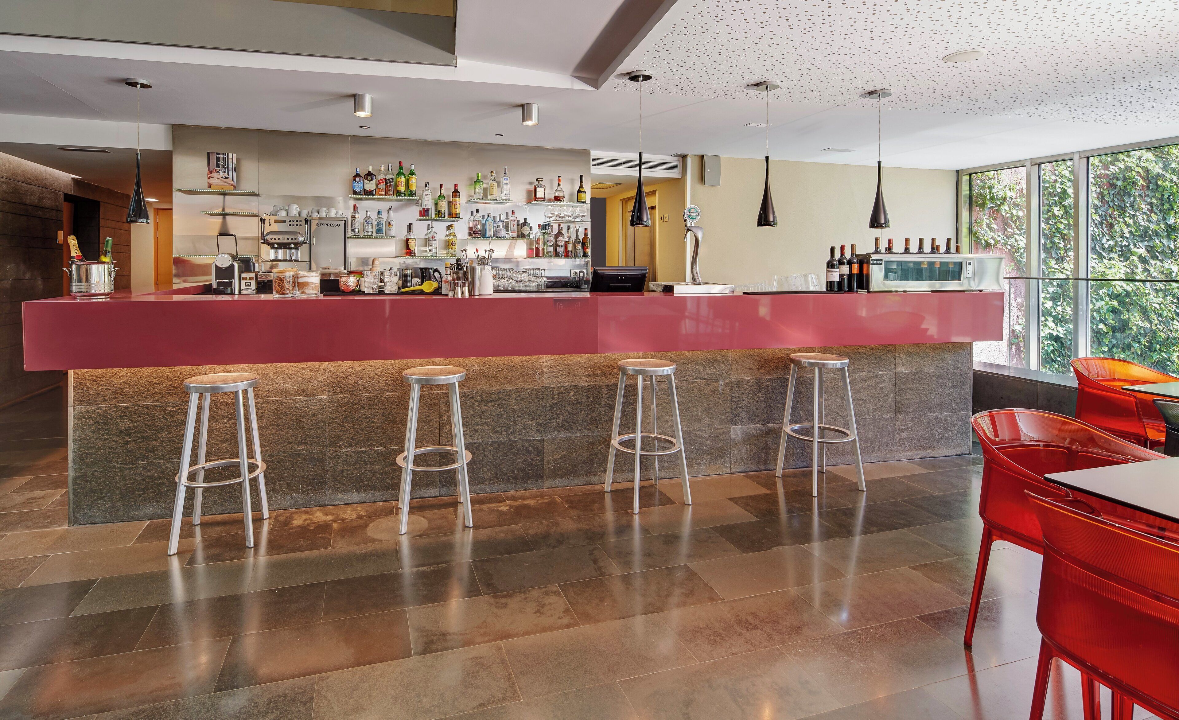 Uma área de bar moderna com balcão vermelho, vários taburetes e uma variedade de garrafas e copos.
