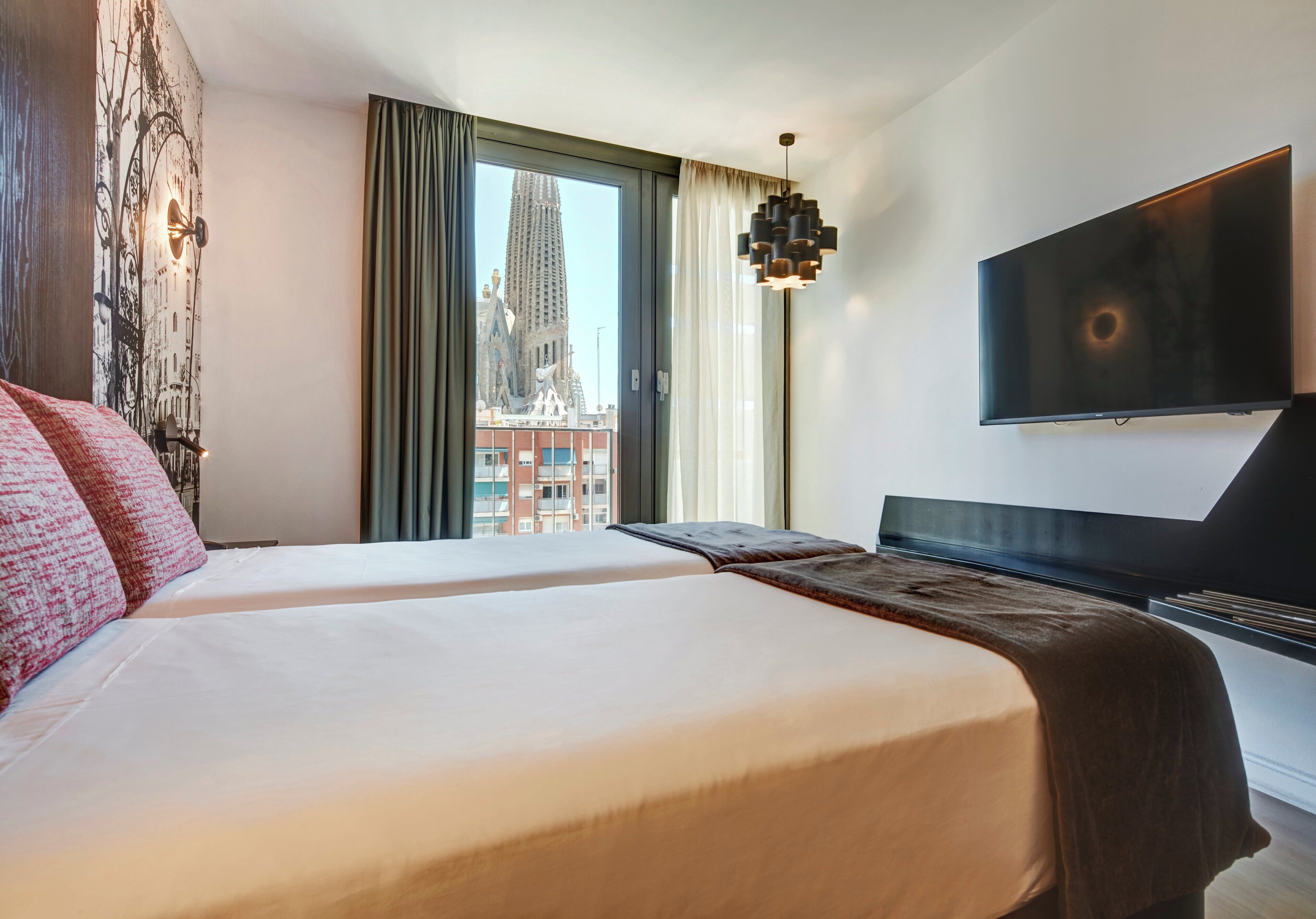 A hotel room with two beds, a large window with a view of a building, a flat-screen TV, and a modern pendant light.