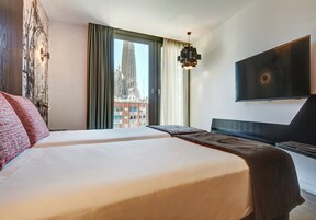 A hotel room with two beds, a large window with a view of a building, a flat-screen TV, and a modern pendant light.