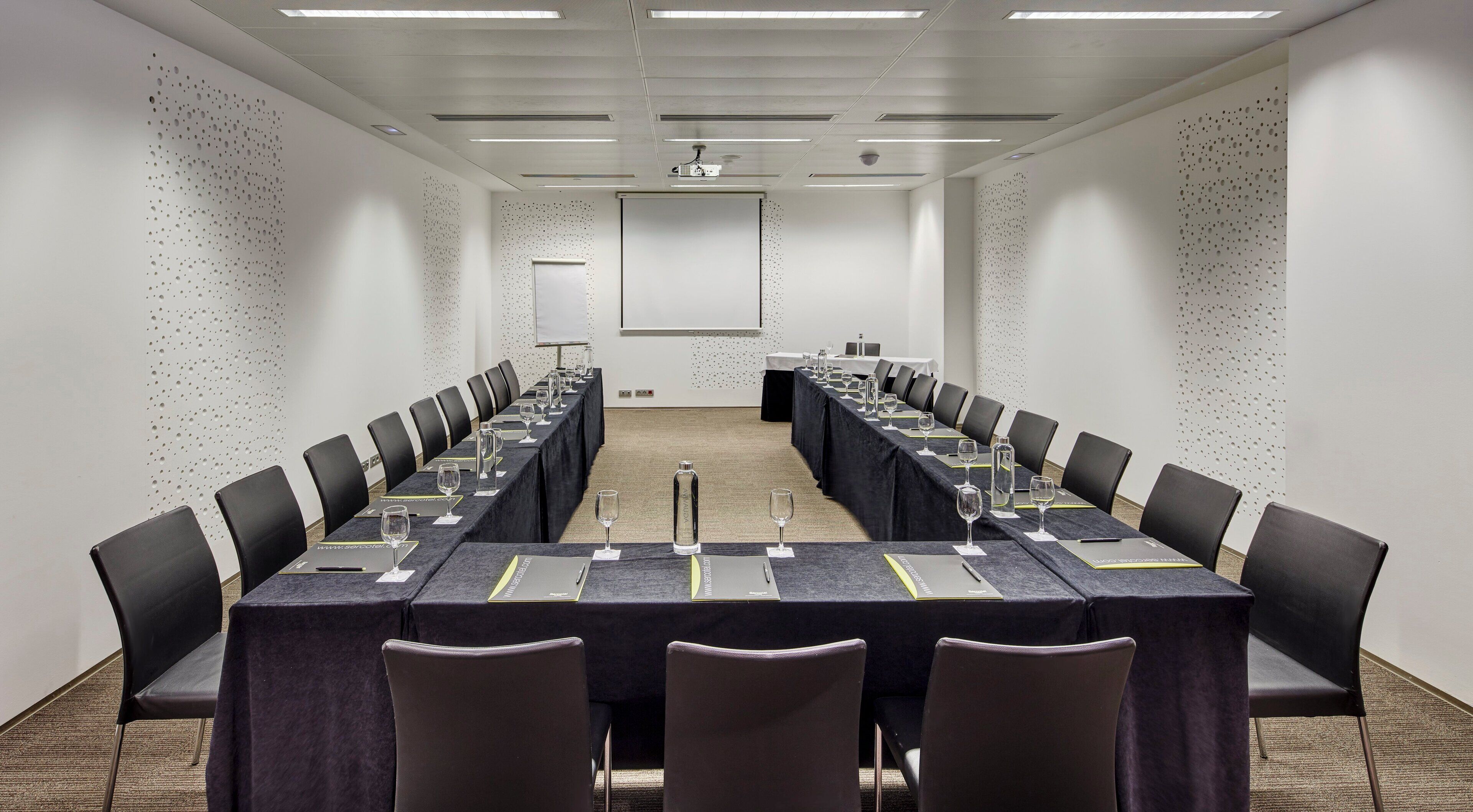 Un salón de conferencias con una larga mesa, sillas negras y una pantalla de proyector.