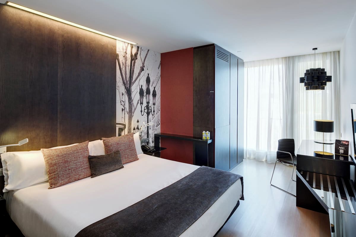 A modern hotel room with a large bed, a desk with a lamp, and a view of the city through the window.