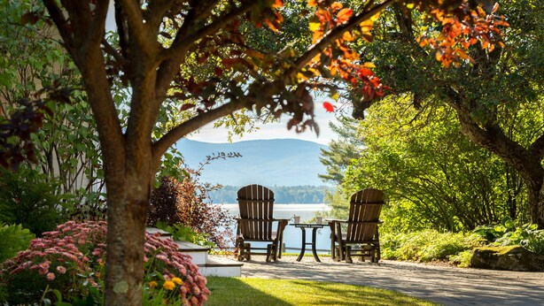 A serene outdoor seating area with two Adirondack chairs and a small table, overlooking a lake and distant mountains. - Inn at Pleasant Lake (New London)