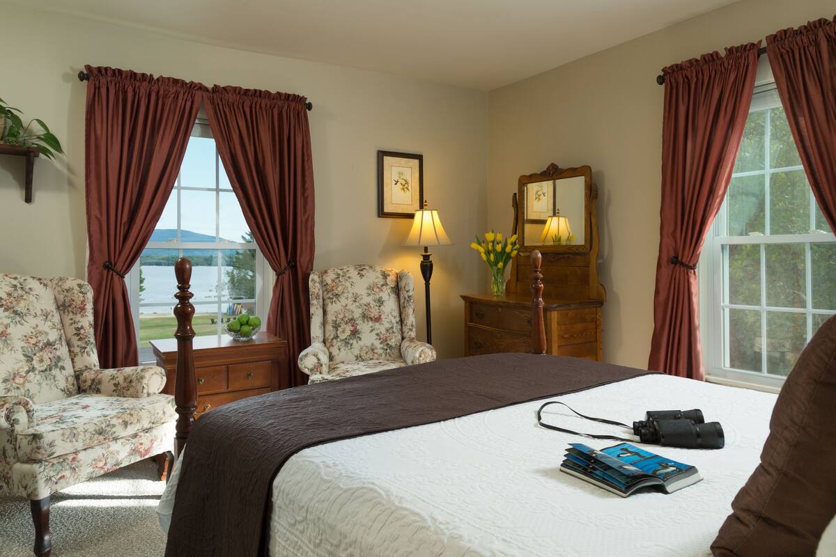 A bedroom with a large bed, two floral armchairs, a wooden dresser, a lamp, and a view of the outdoors.