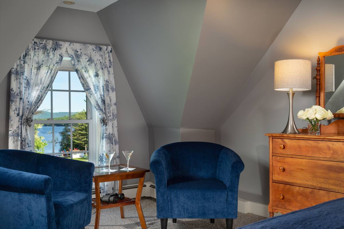 A room with a view of a lake, a wooden dresser, a blue armchair, a table with a glass, and a lamp.