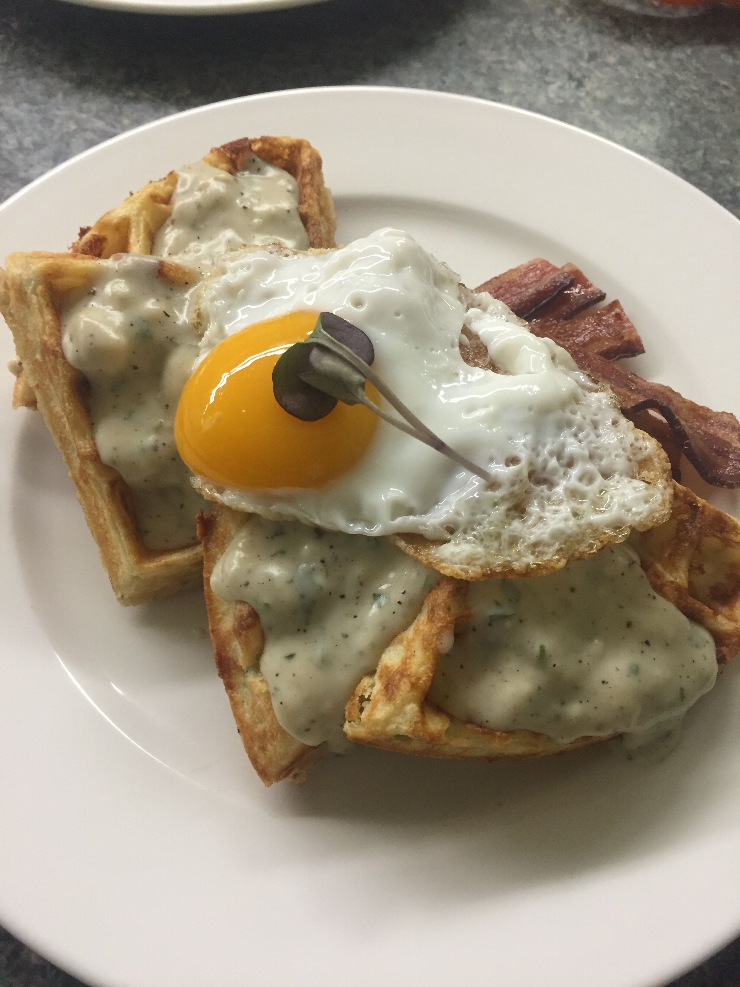 Une assiette avec un œuf au plat sur une tranche de pain grillé, nappée d’une sauce crémeuse, et accompagnée de lardons.