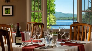 A dining table set with wine glasses, a bottle of Tres Picos wine, and a view of a lake through large windows. - Inn at Pleasant Lake (New London)