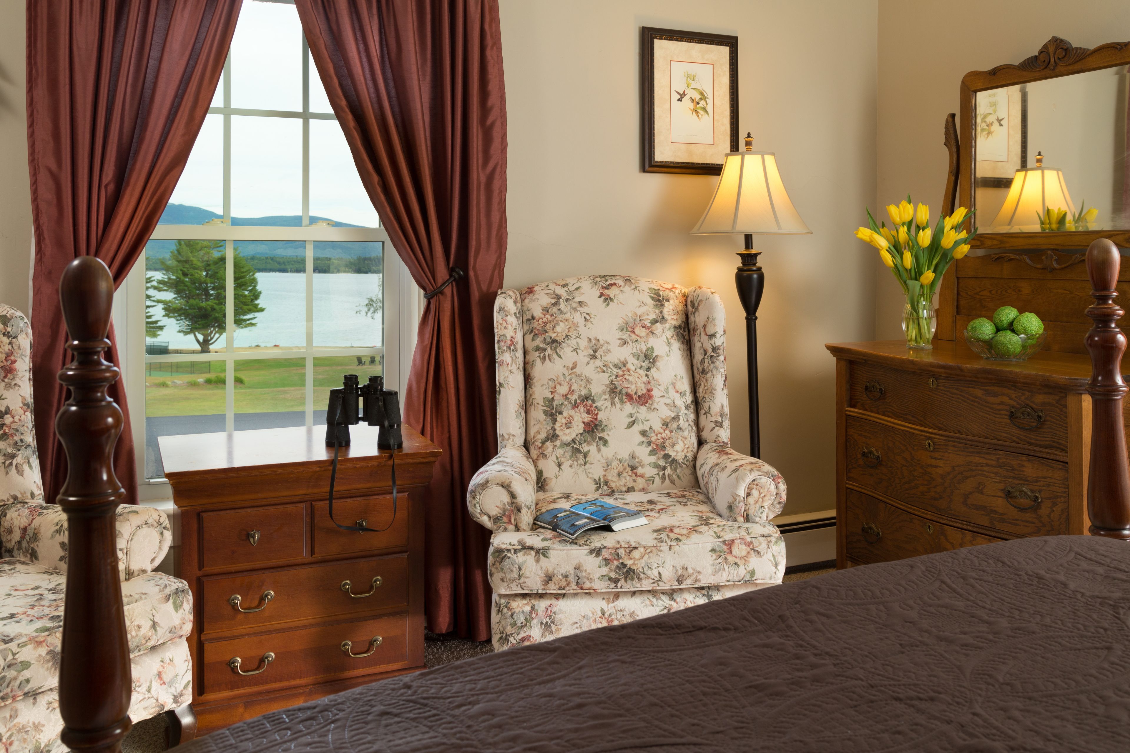 Un dormitorio con una cama, un sillón floral, un aparador de madera, una lámpara y un ventanal con cortinas.