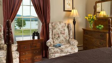 Un dormitorio con una cama, un sillón floral, un aparador de madera, una lámpara y un ventanal con cortinas.