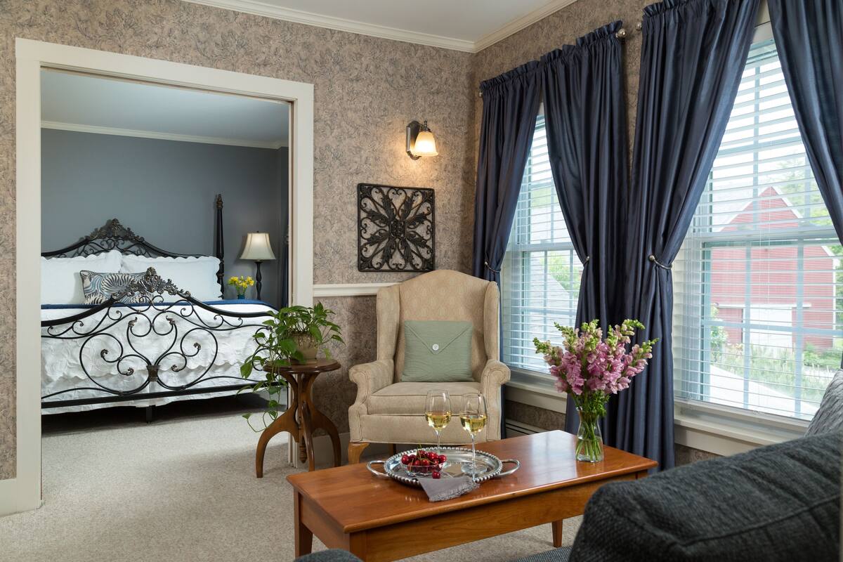 A bedroom with a bed, a chair, a table with a tray of fruit and glasses, and a window with curtains.