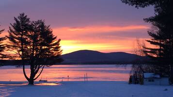 A snowy landscape with a sunset over a body of water, trees, and a distant mountain.