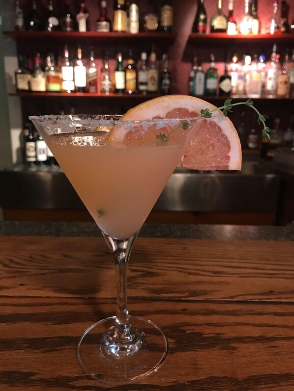 A cocktail in a martini glass with a citrus garnish on a wooden bar counter.