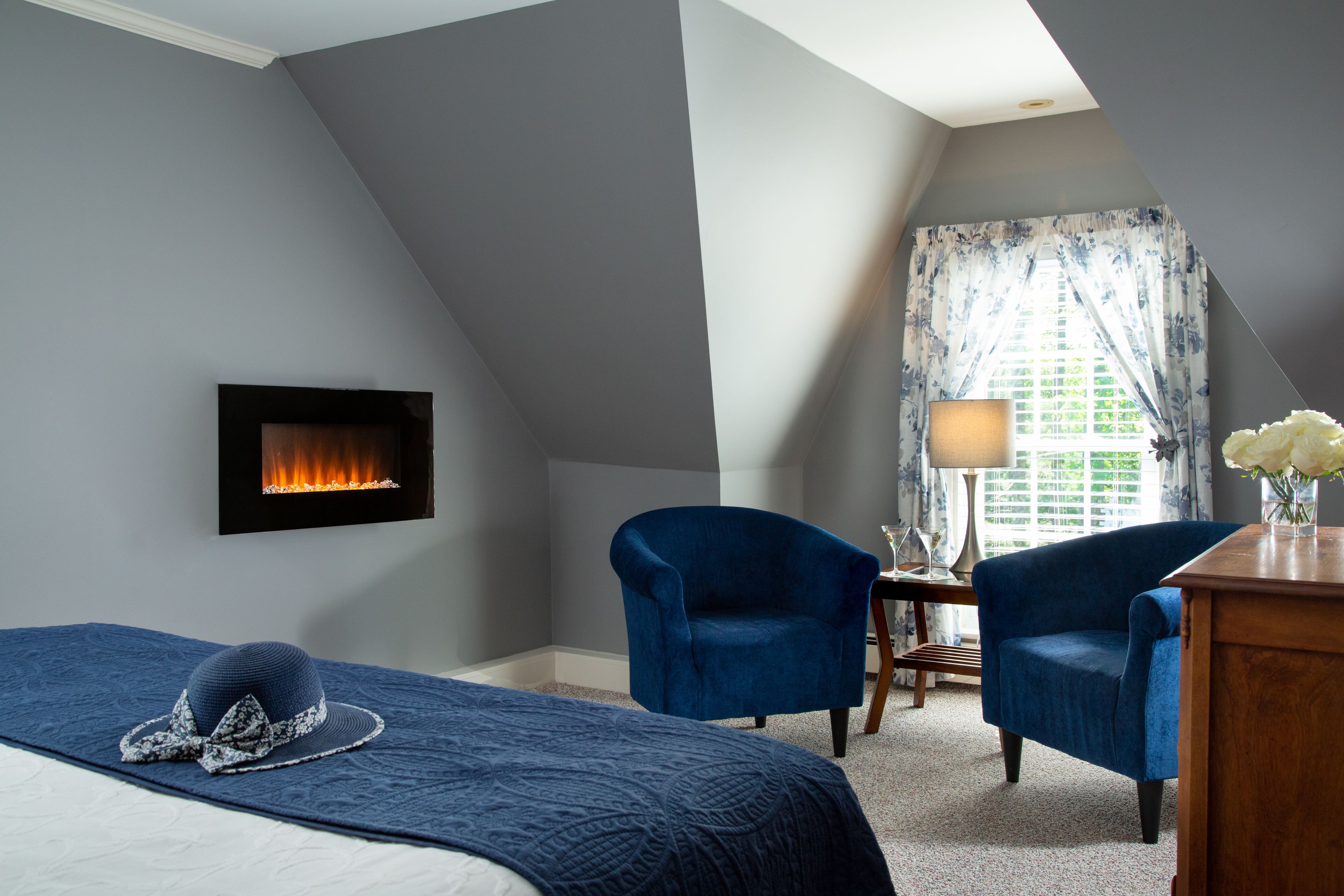 Un dormitorio con chimenea, dos sillones azules, una cama con colcha azul y una ventana con cortinas florales.