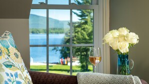 A window with a view of a lake and mountains, a vase with white roses, and a glass of wine on a table. - Inn at Pleasant Lake (New London)