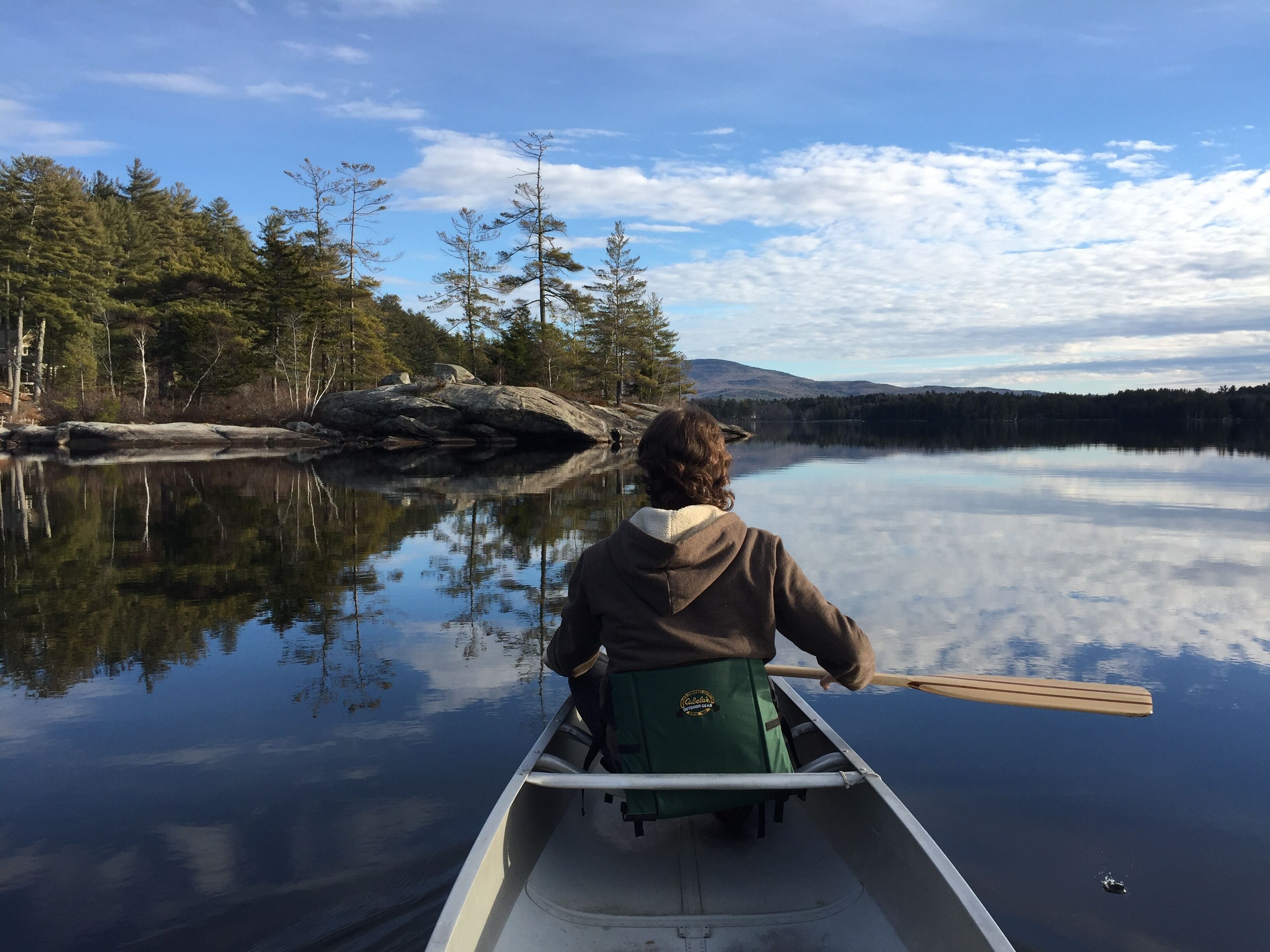 A person in a canoe on a calm lake with trees and mountains in the background.