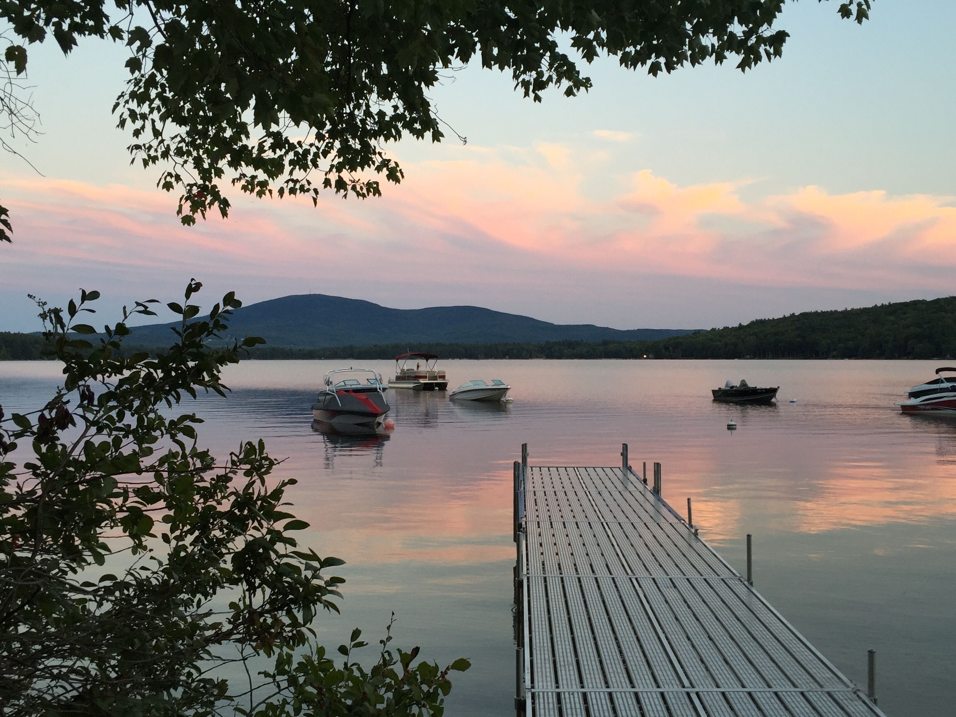 A calm lake with boats docked at a wooden pier, surrounded by trees and hills in the background.