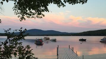 A calm lake with boats docked at a wooden pier, surrounded by trees and hills in the background.