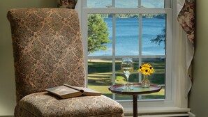A room with a large window offering a view of a lake and hills. A chair with a book and a glass of wine on a small table beside the window. - Inn at Pleasant Lake (New London)