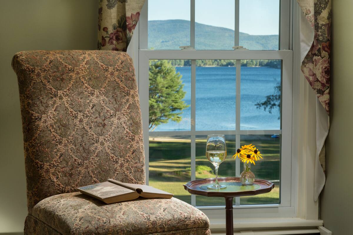 A room with a large window offering a view of a lake and hills. A chair with a book and a glass of wine on a small table beside the window.