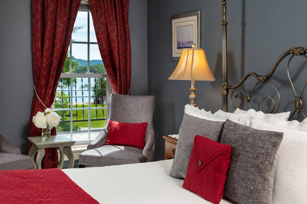 A bedroom with a bed, two chairs, a nightstand, a lamp, and a window with curtains.